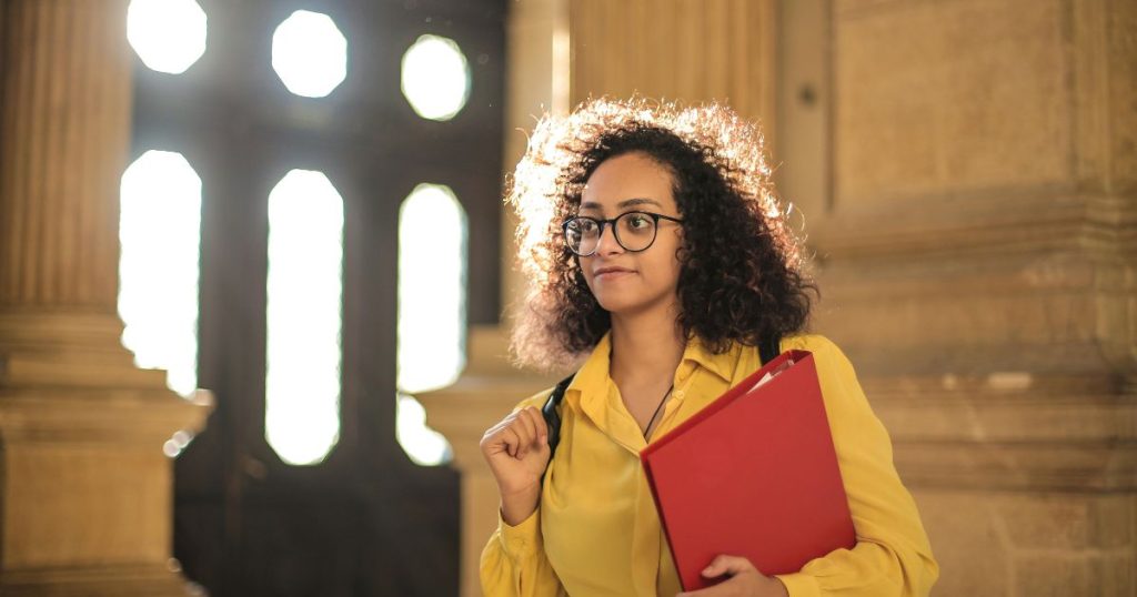 A confident student in a yellow shirt holding a red folder, standing in a grand hallway, representing a recipient of the GGU Worldwide Scholarship (UHUB).